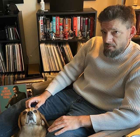 professor stephen cummings, pictured in a white sweater and jeans next to his beagle and his collection of vivnyl records