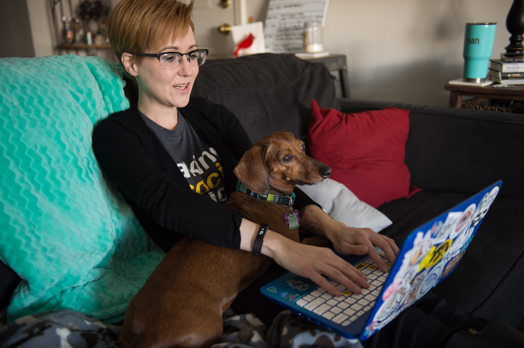 a white woman with short hair and glasses sitting on her couch with her laptop computer and her wiener dog on her lap