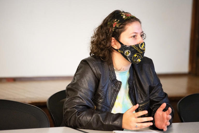 Female student seated in class wearing a black leather jacket and face mask, gesturing with her hands