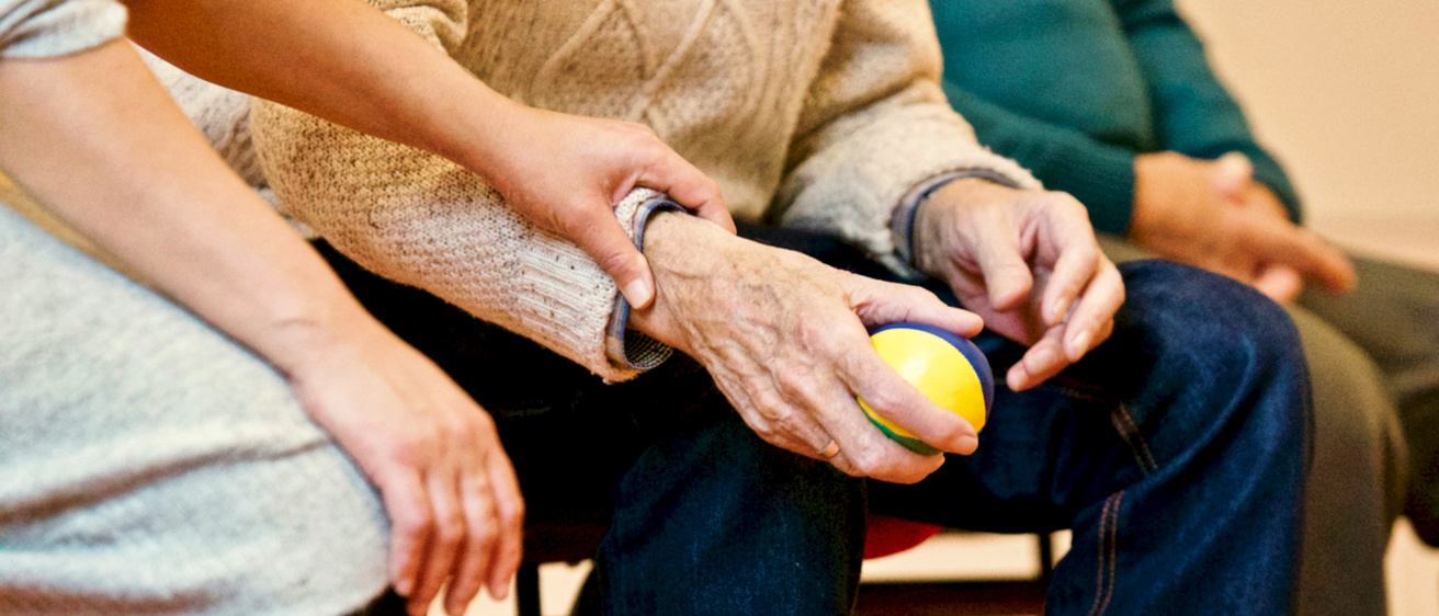 Elderly man holding a stress ball, assisted by the hand of a younger person.