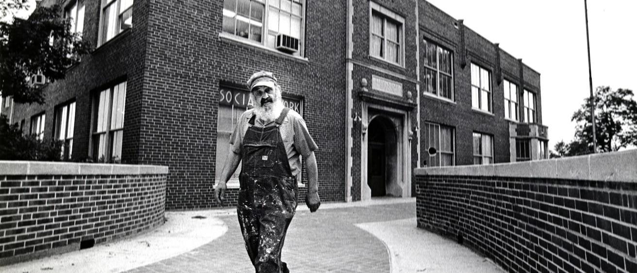black and white image of Bill sackter, wearing overalls and an engineer cap, walking outside North Hall
