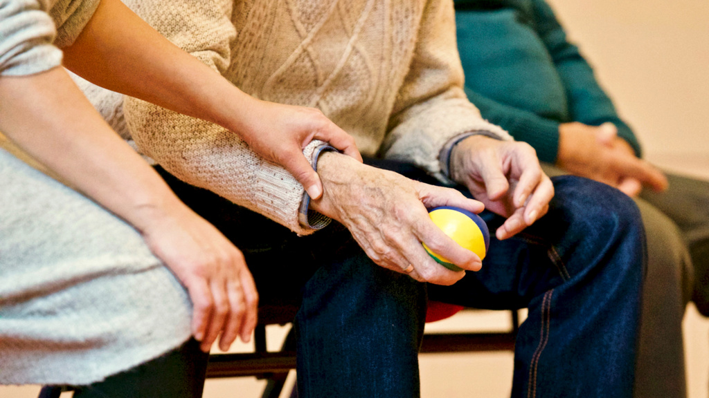 Elderly man holding a stress ball, assisted by the hand of a younger person.