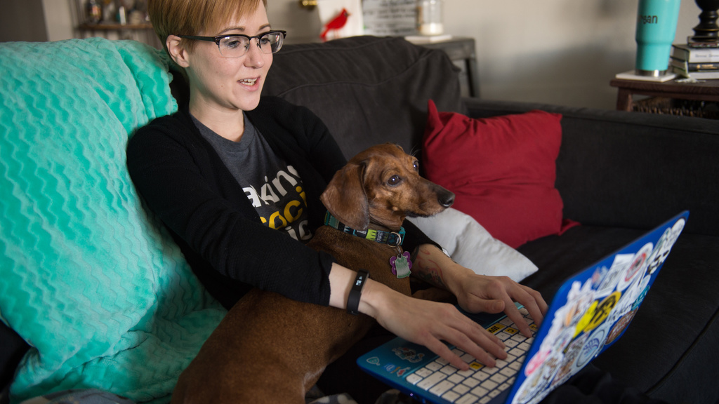 a white woman with short hair and glasses sitting on her couch with her laptop computer and her wiener dog on her lap